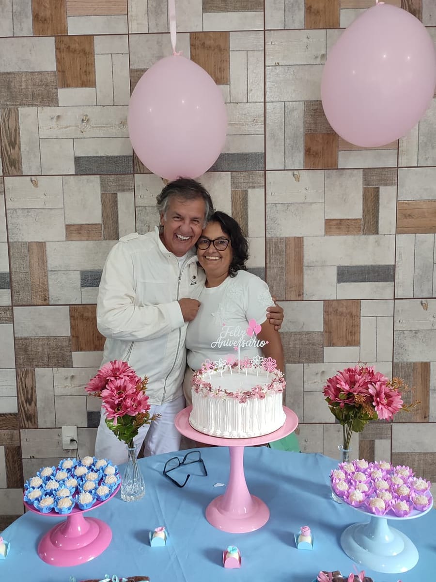 Homem abraça uma mulher atrás de uma mesa de bolo de aniversário decorada em rosa.