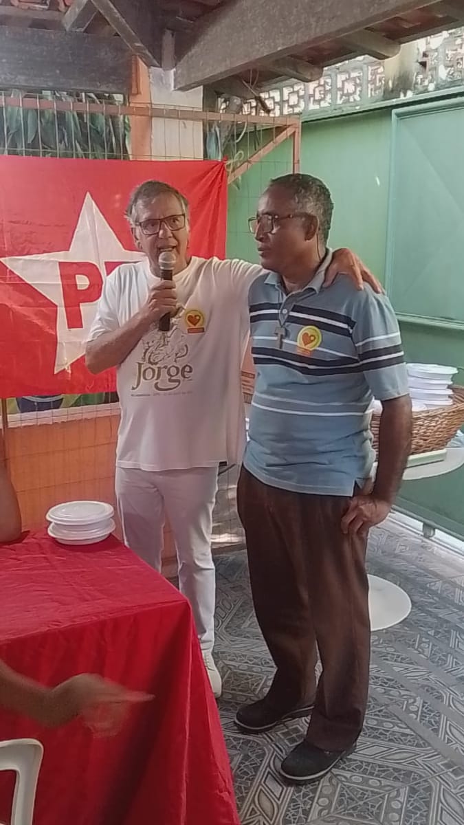 Homem ao microfone abraça pelo ombro um companheiro diante da bandeira do partido.