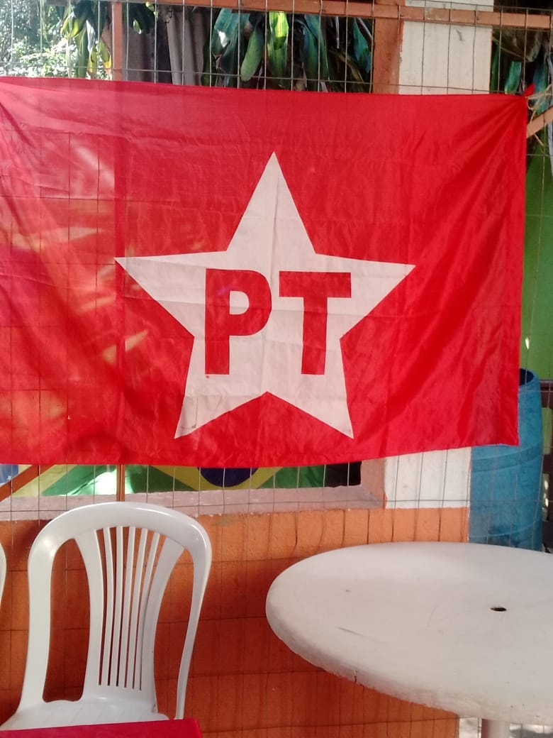 Bandeira vermelha do partido estendida na parede de um salão, ao lado de mesa e cadeiras de plástico.