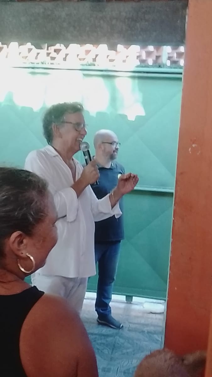 Homem fala ao microfone enquanto pessoas o escutam de perto em um salão comunitário.