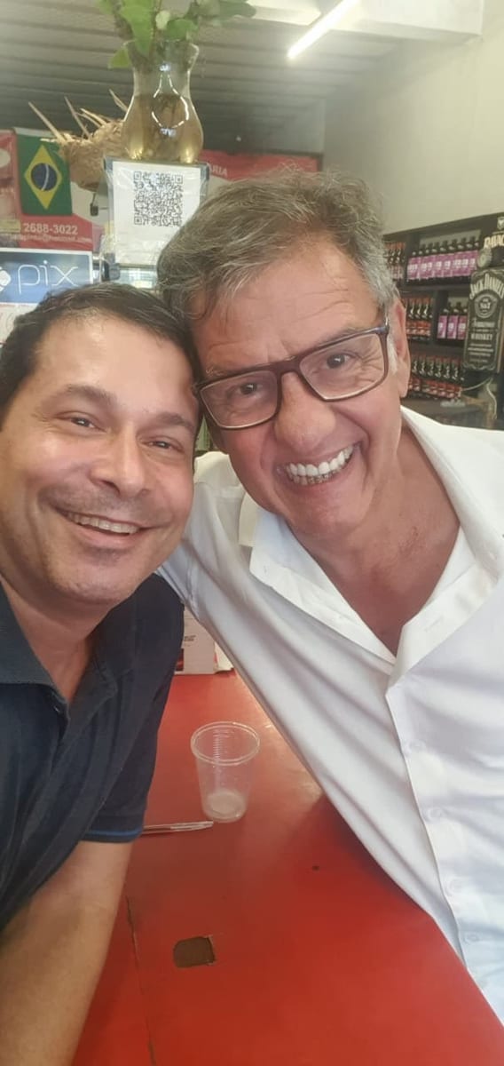 Dois homens sorriem lado a lado em uma mesa de estabelecimento comercial.