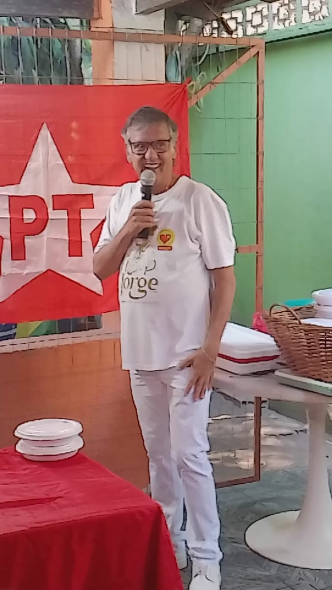 Homem de branco ri enquanto fala ao microfone diante da bandeira do partido.
