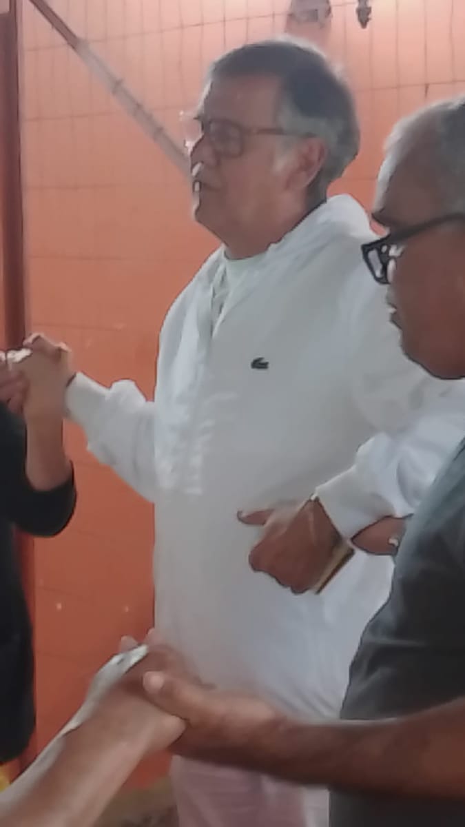 Homem de camisa branca aperta as mãos de pessoas ao seu redor em um salão comunitário.