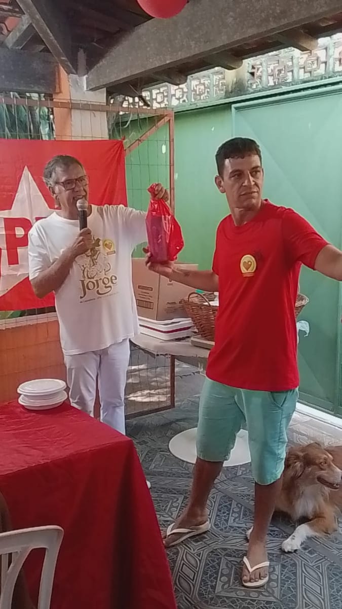 Dr. Marquinho Barreto, ao microfone, entrega uma sacola vermelha a um voluntário durante uma atividade comunitária.