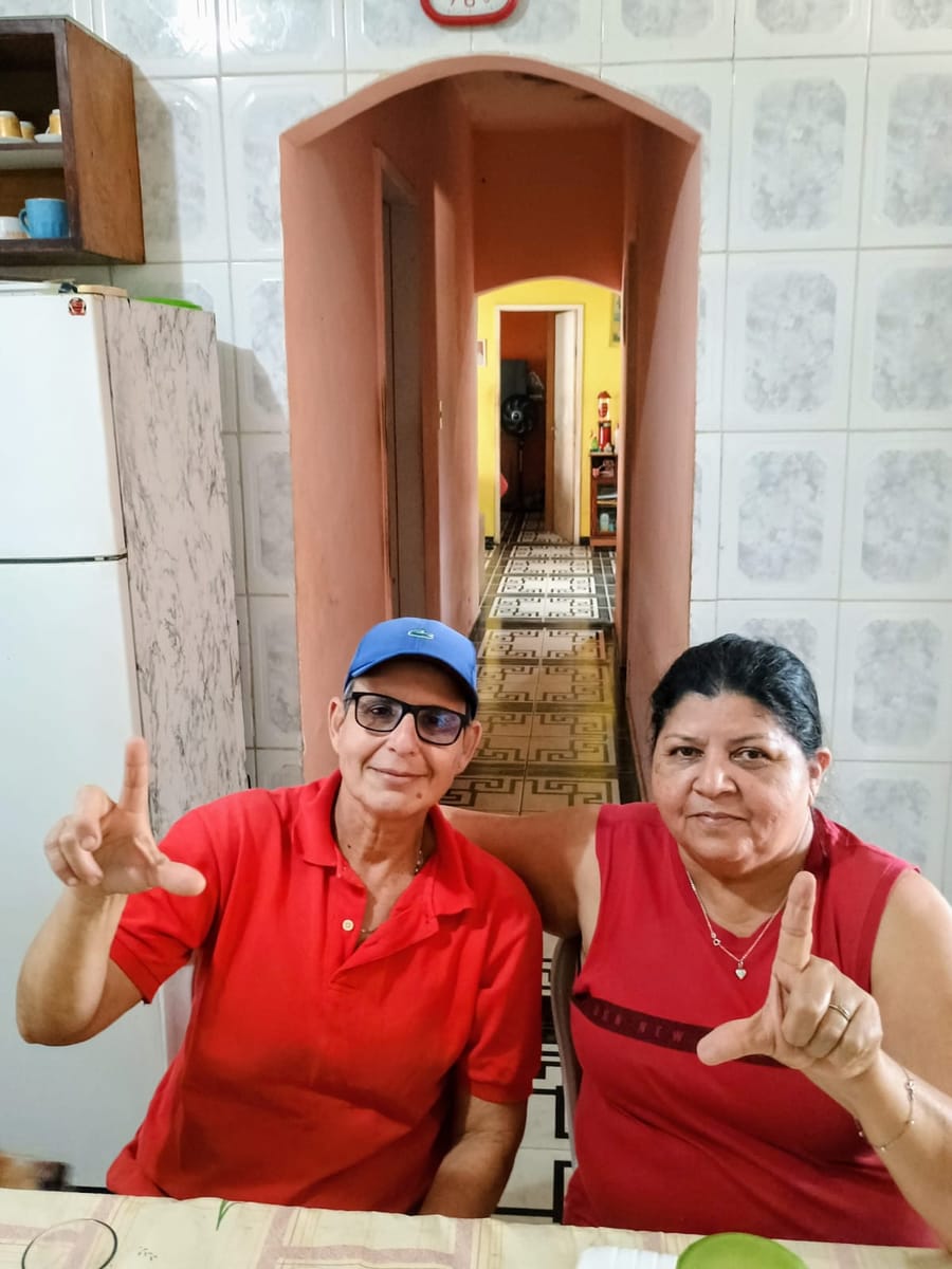 Duas pessoas sentadas à mesa de uma cozinha erguem os dedos formando a letra L.