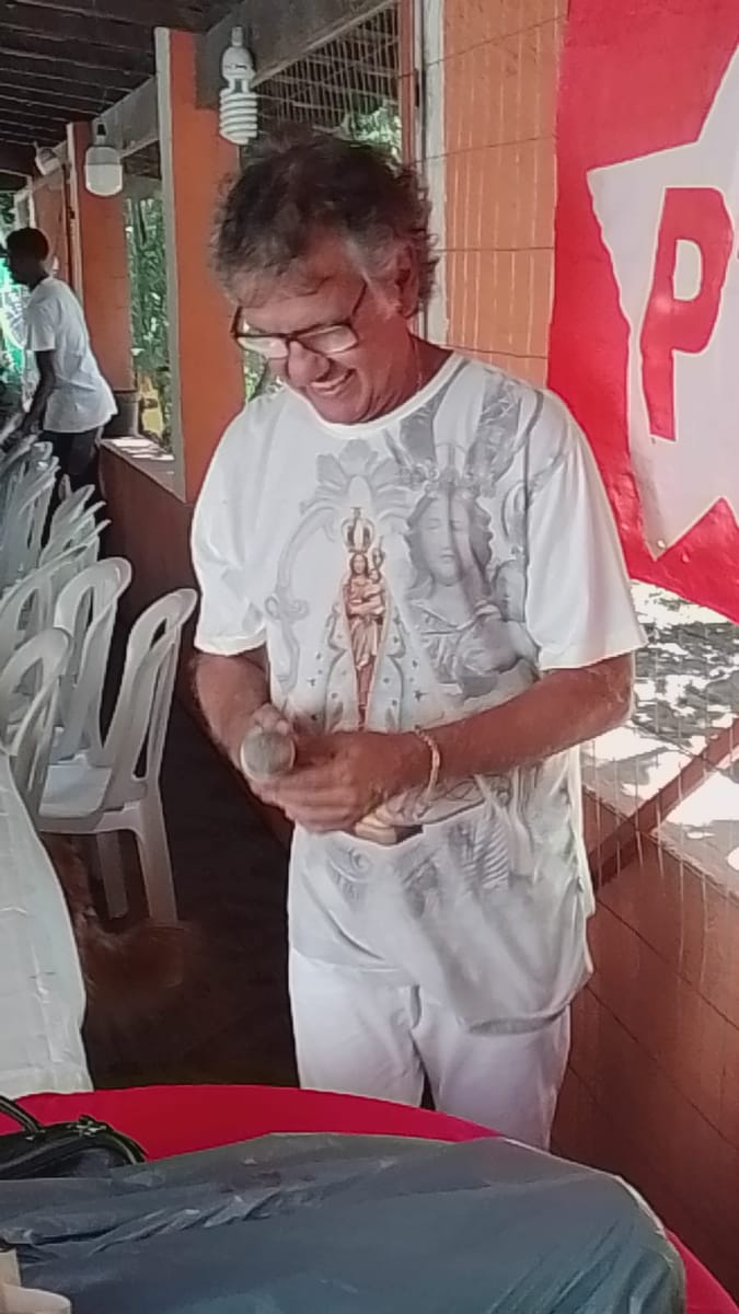 Homem segura com cuidado uma pequena imagem de santo em um salão decorado com bandeira do partido.