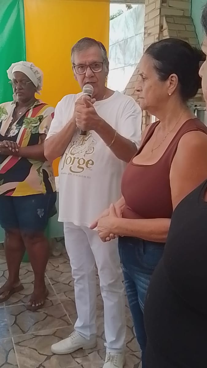 Dr. Marquinho Barreto fala ao microfone rodeado de mulheres que o escutam de perto em um salão comunitário.