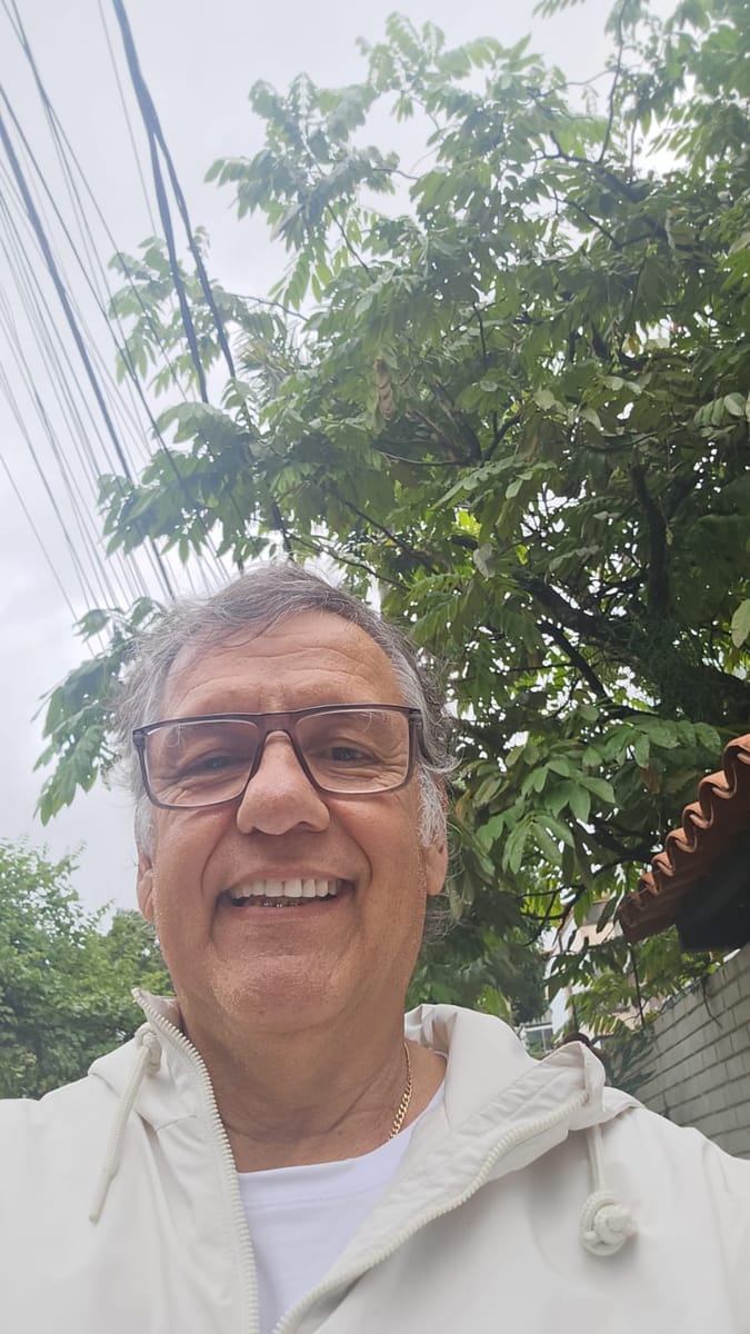Homem de cabelos grisalhos e óculos sorri ao ar livre, com a copa de uma árvore atrás dele.