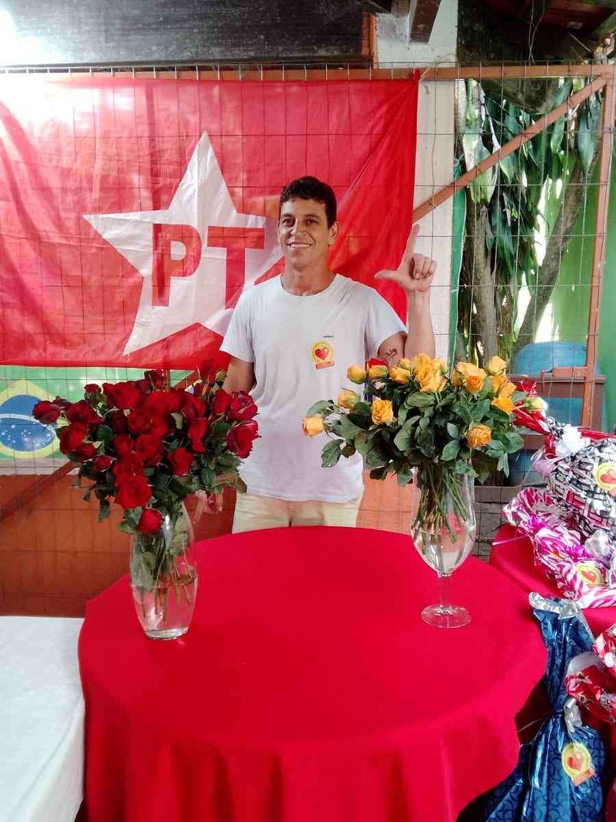 Homem sorri e ergue os dedos formando a letra L diante de uma bandeira do partido, entre vasos de rosas.