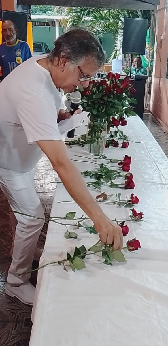Homem de branco arruma rosas vermelhas, uma a uma, sobre uma mesa comprida coberta de toalha branca.