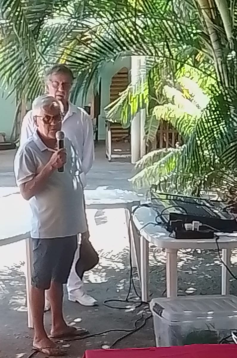Um senhor fala ao microfone ao ar livre enquanto outro homem aguarda sua vez atrás dele.