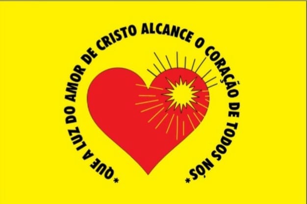 Bandeira amarela com um coração vermelho e um sol irradiando ao centro, cercado pela frase &ldquo;Que a luz do amor de Cristo alcance o coração de todos nós&rdquo;.