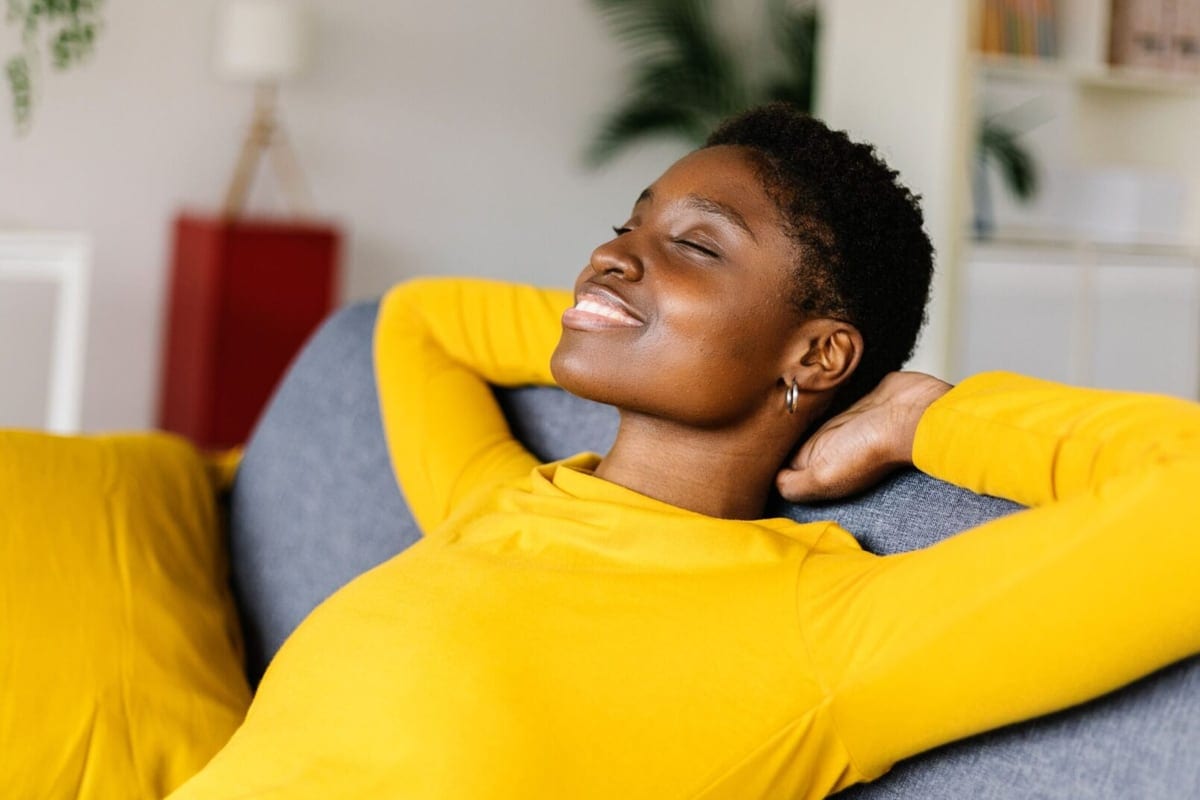 Mulher de blusa amarela descansa recostada no sofá, de olhos fechados e sorrindo.