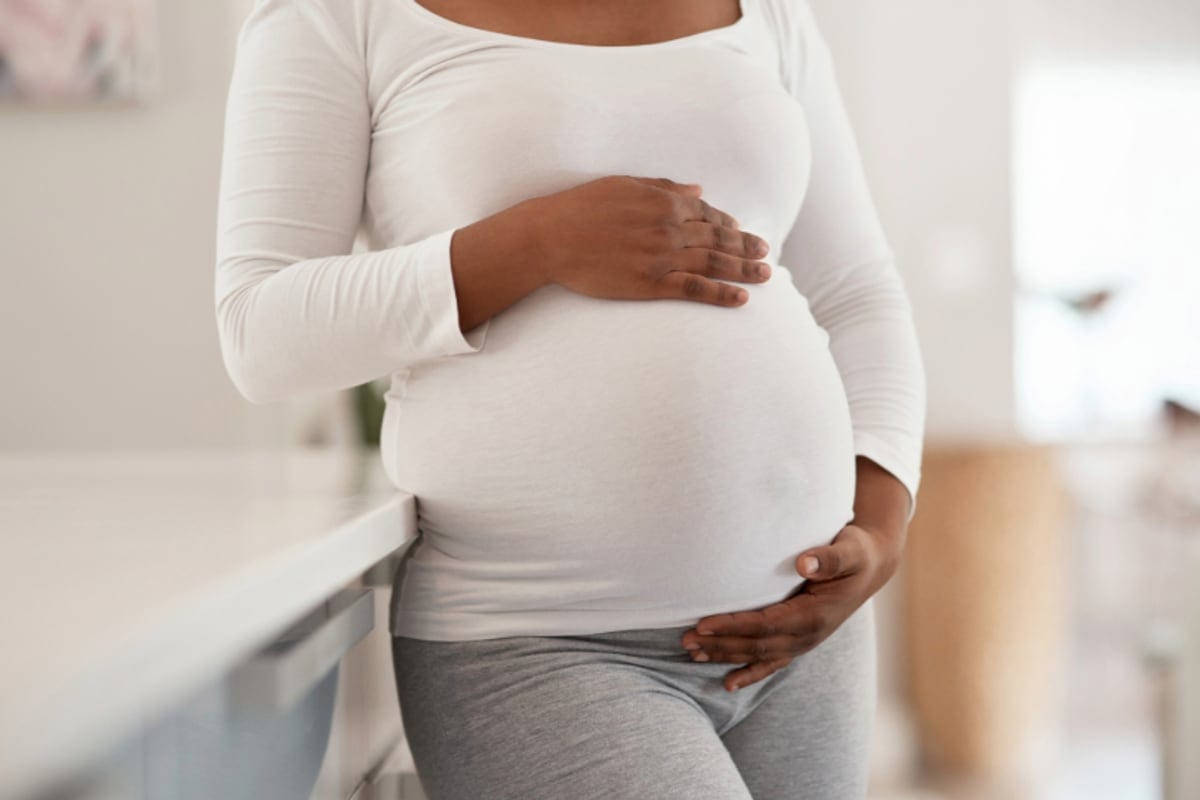 Mulher grávida em pé, com as duas mãos apoiadas sobre a barriga. O rosto está fora do quadro.