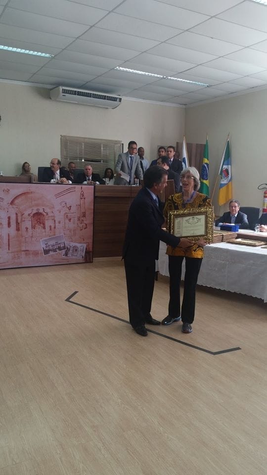 Entrega de um título honorífico no plenário da Câmara, com a mesa diretora e bandeiras ao fundo.