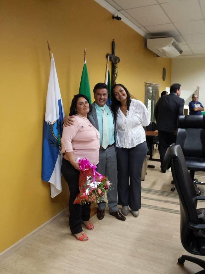 Três pessoas posam juntas na Câmara Municipal diante das bandeiras de Itaguaí, do Brasil e do Rio de Janeiro.