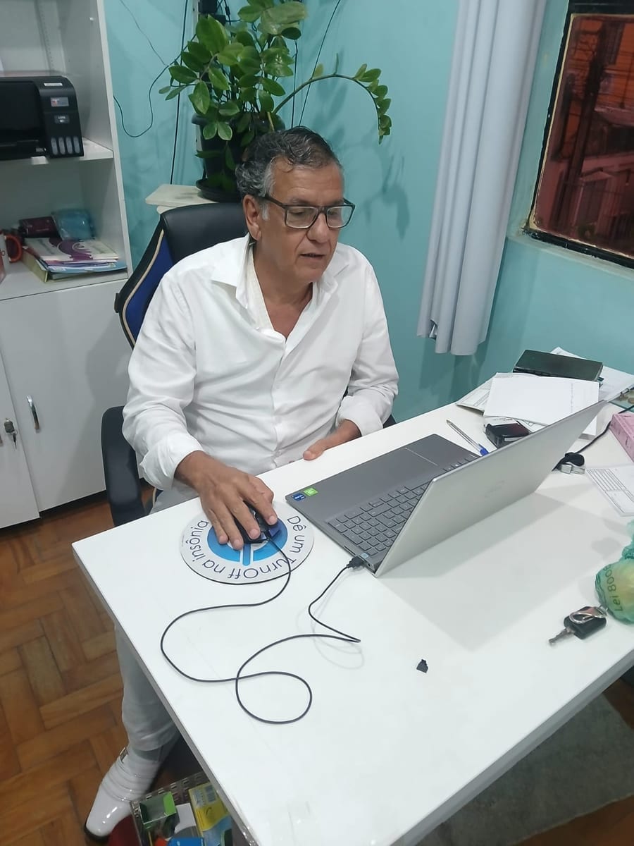 Dr. Marquinho Barreto sentado à mesa de trabalho, de camisa branca, diante de um notebook.