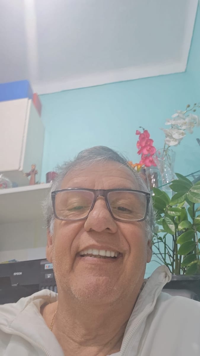 Dr. Marquinho Barreto de óculos sorri em uma sala clara, com orquídeas ao fundo.