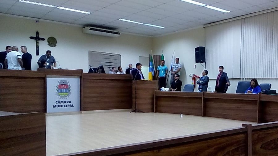 Plenário da Câmara Municipal de Itaguaí em plano aberto, com brasão e bandeiras ao fundo.