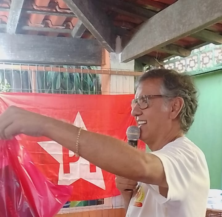 Dr. Marquinho Barreto, de perfil e com microfone na mão, durante um encontro; ao fundo, uma bandeira do PT.