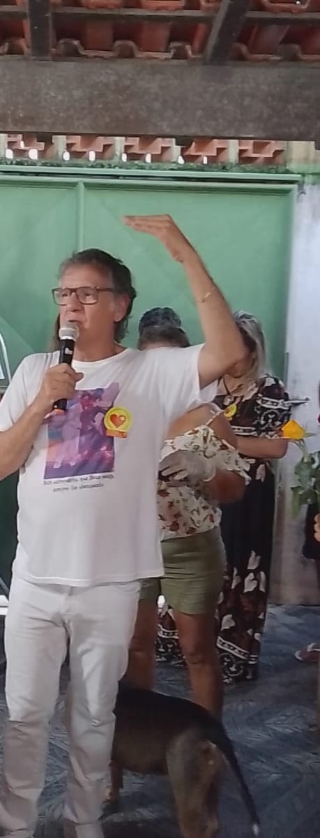 Dr. Marquinho Barreto fala ao microfone, de camiseta branca com um coração estampado, cercado por apoiadoras que erguem as mãos.