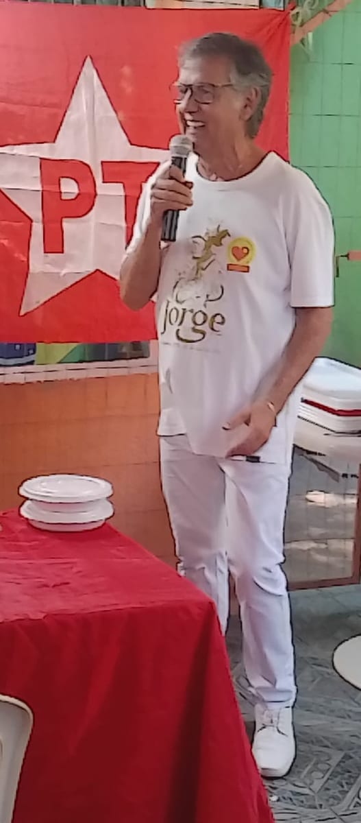 Dr. Marquinho Barreto fala ao microfone, em pé, diante de uma bandeira do PT, num quintal coberto.
