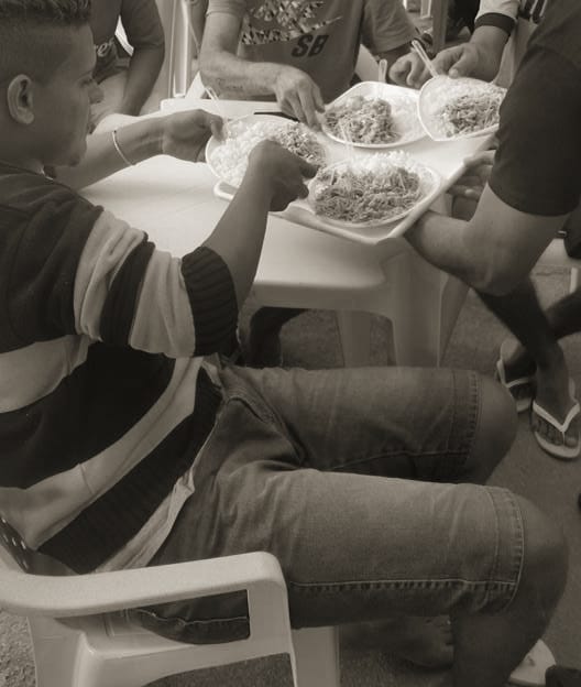 Rapazes servindo-se de bandejas com arroz e macarrão durante um almoço comunitário.