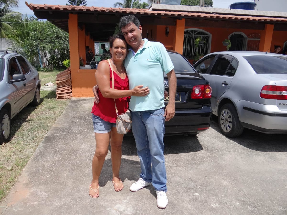 Homem e mulher posam abraçados e sorrindo em frente a uma casa de fachada laranja.