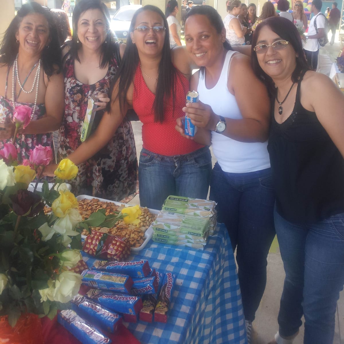 Cinco mulheres sorriem abraçadas atrás de uma mesa com rosas e guloseimas.