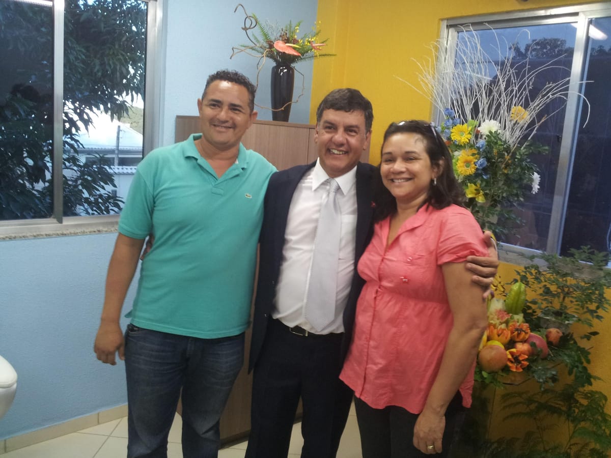 Três adultos posam abraçados e sorrindo em uma sala decorada com arranjos florais.