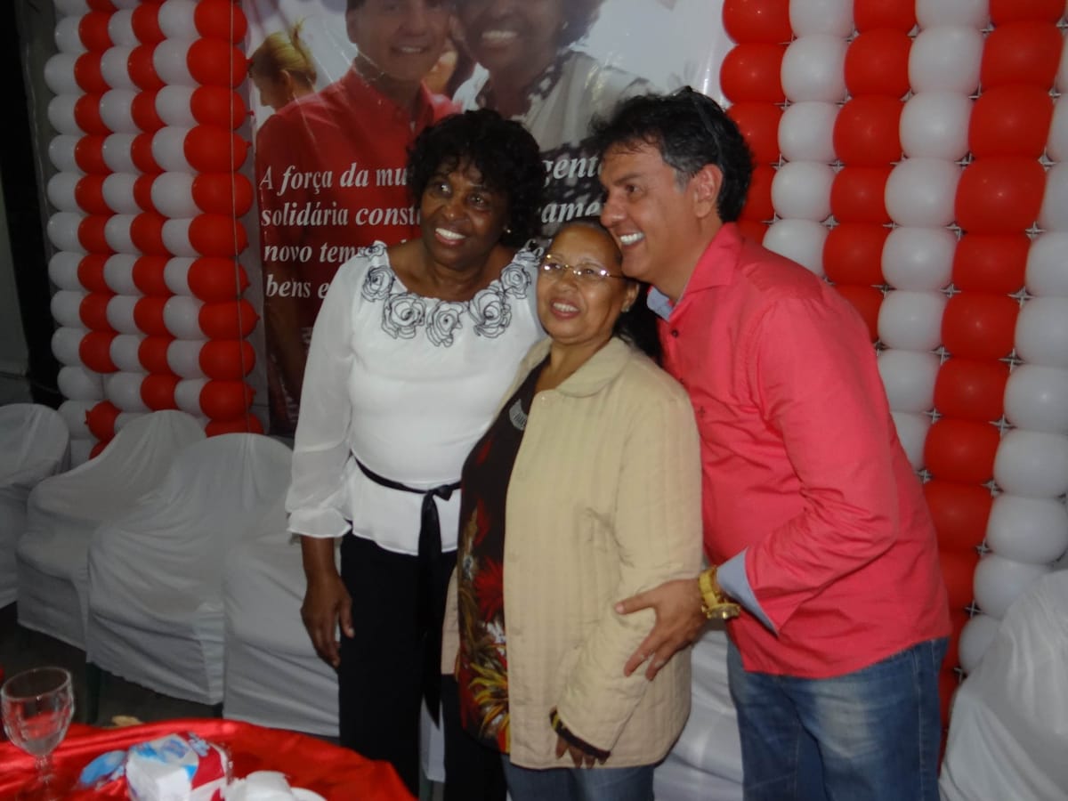 Homem posa sorrindo ao lado de duas senhoras diante de uma faixa e de colunas de balões vermelhos e brancos.