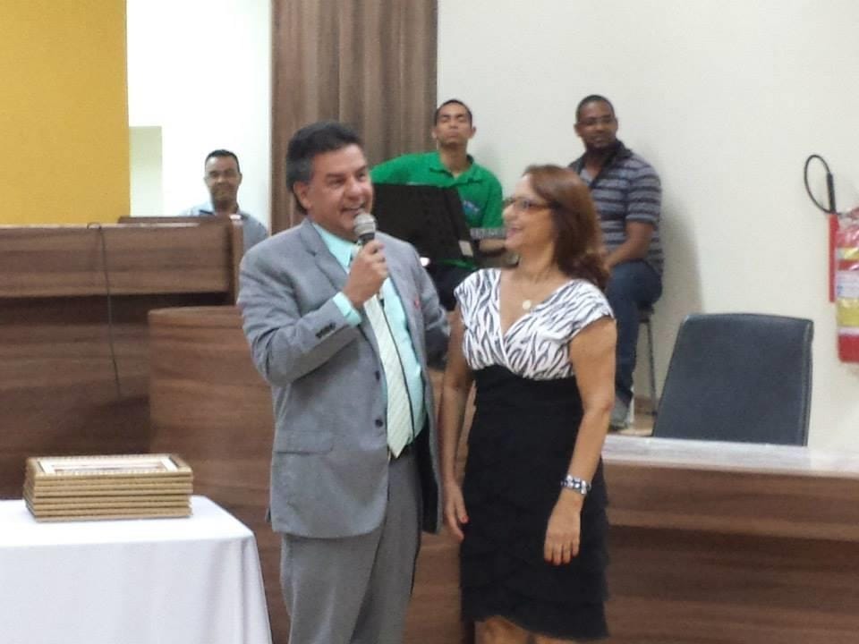 Homem de terno fala ao microfone ao lado de uma mulher no plenário de uma câmara municipal.