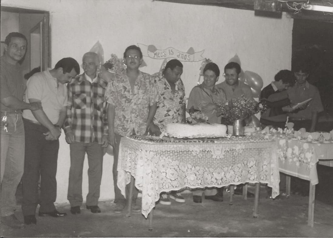 Nove homens adultos posam atrás de uma mesa com bolo, sob uma faixa artesanal escrita “Meus 15 anos”.