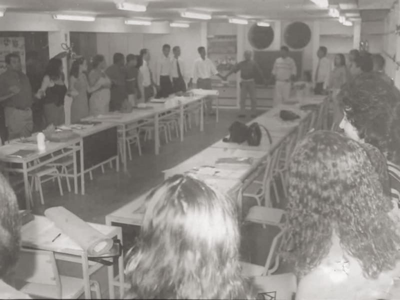 Sala de aula com carteiras amarelas onde um grupo de adultos formalmente vestidos está de mãos dadas em fila.