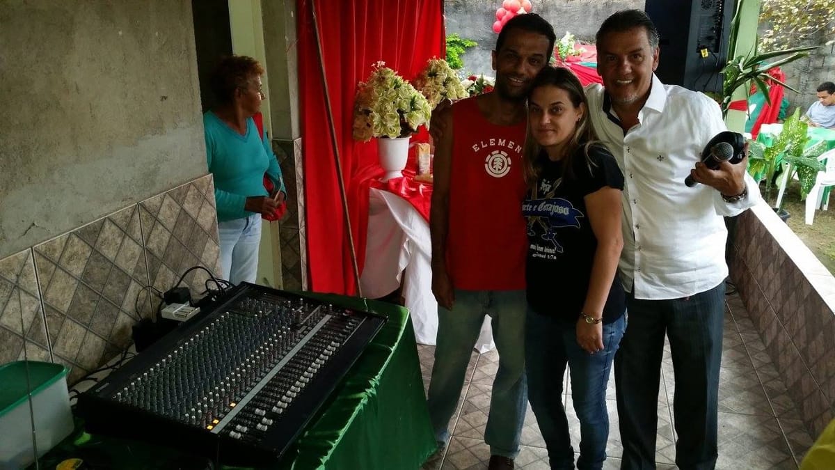 Homem segurando um microfone posa ao lado de um casal, junto a uma mesa de som em festa comunitária.