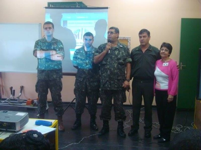 Três militares fardados, um deles falando ao microfone, posam ao lado de um homem e uma mulher diante de uma tela de projeção.