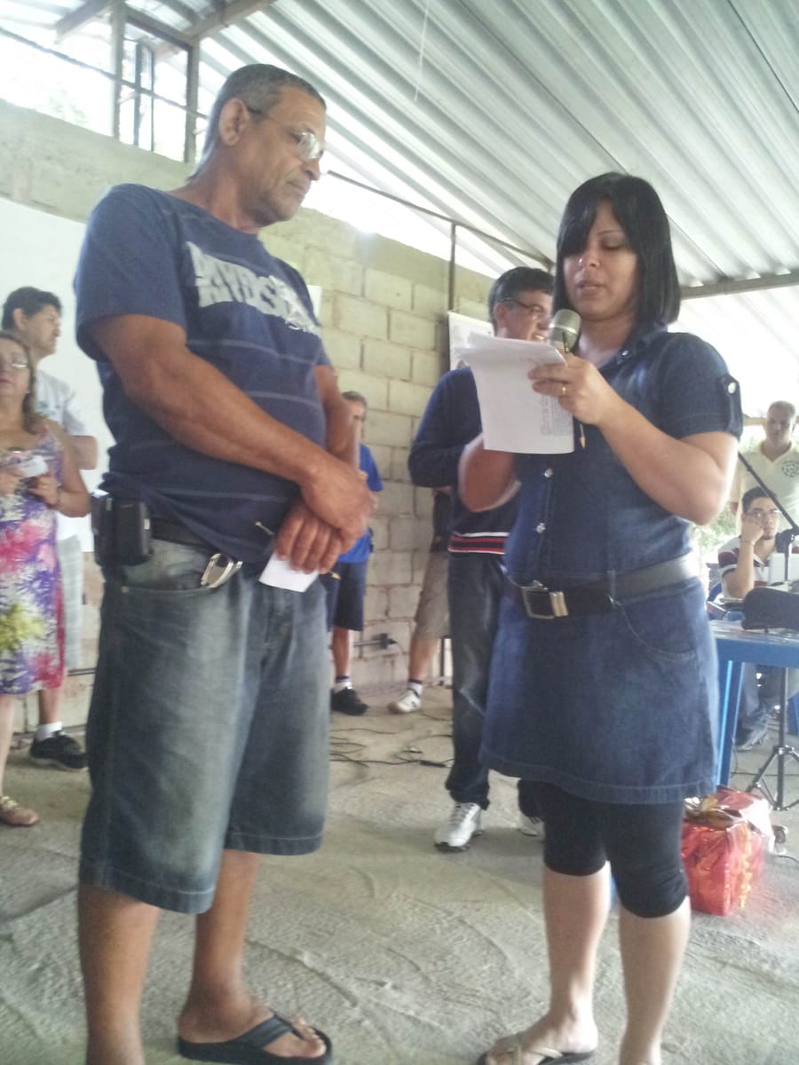 Mulher lê um documento ao microfone ao lado de um homem, sob cobertura de zinco, diante de um pequeno grupo.