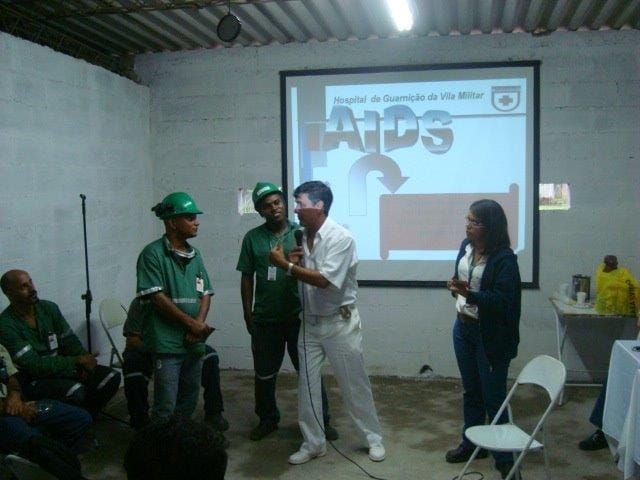 Homem apresenta um slide sobre AIDS a um grupo de trabalhadores uniformizados e de capacete.