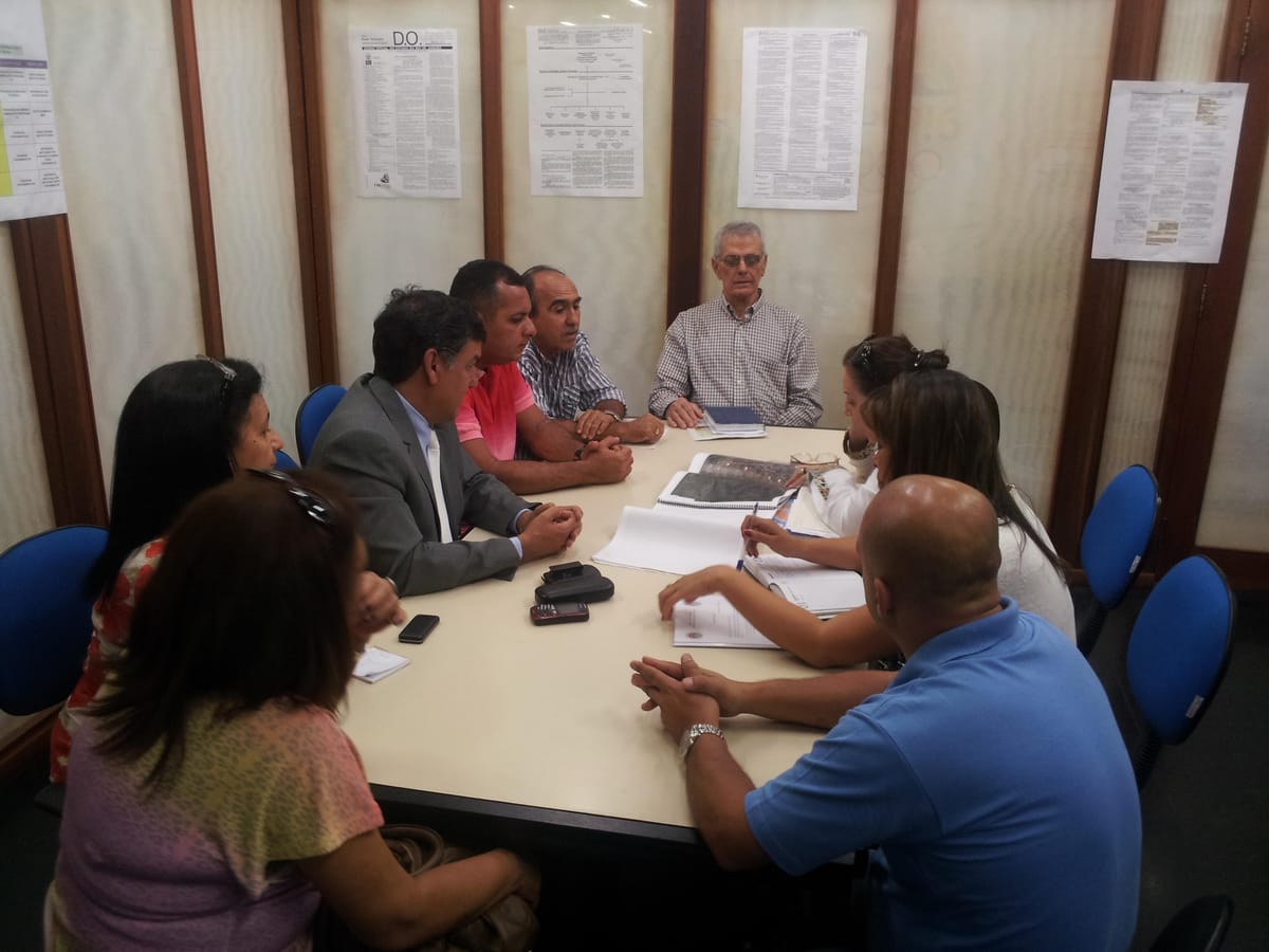 Grupo de adultos reunido em torno de uma mesa de trabalho, com páginas de Diário Oficial afixadas na parede.