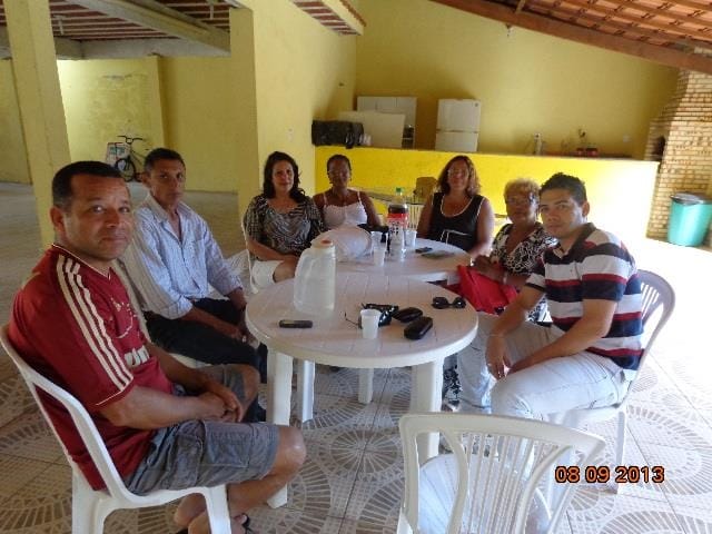 Sete adultos sentados em torno de uma mesa sob uma cobertura simples.