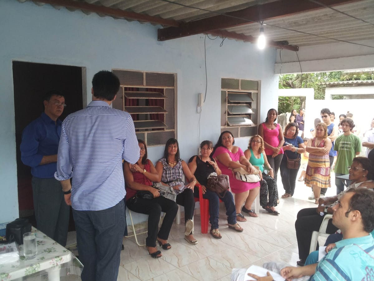 Grupo de moradores sentados em cadeiras na varanda de uma casa, acompanhando dois homens que conversam em pé.