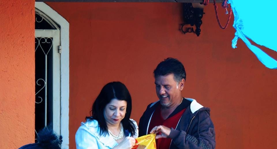 Uma mulher e um homem abrem juntos um saco de balas em frente a uma parede laranja.