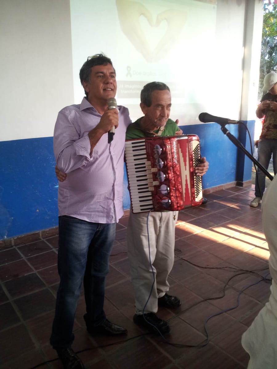 Homem ao microfone abraça um senhor idoso que segura uma sanfona vermelha, diante de uma projeção com a mensagem “Doe órgãos, doe vida”.